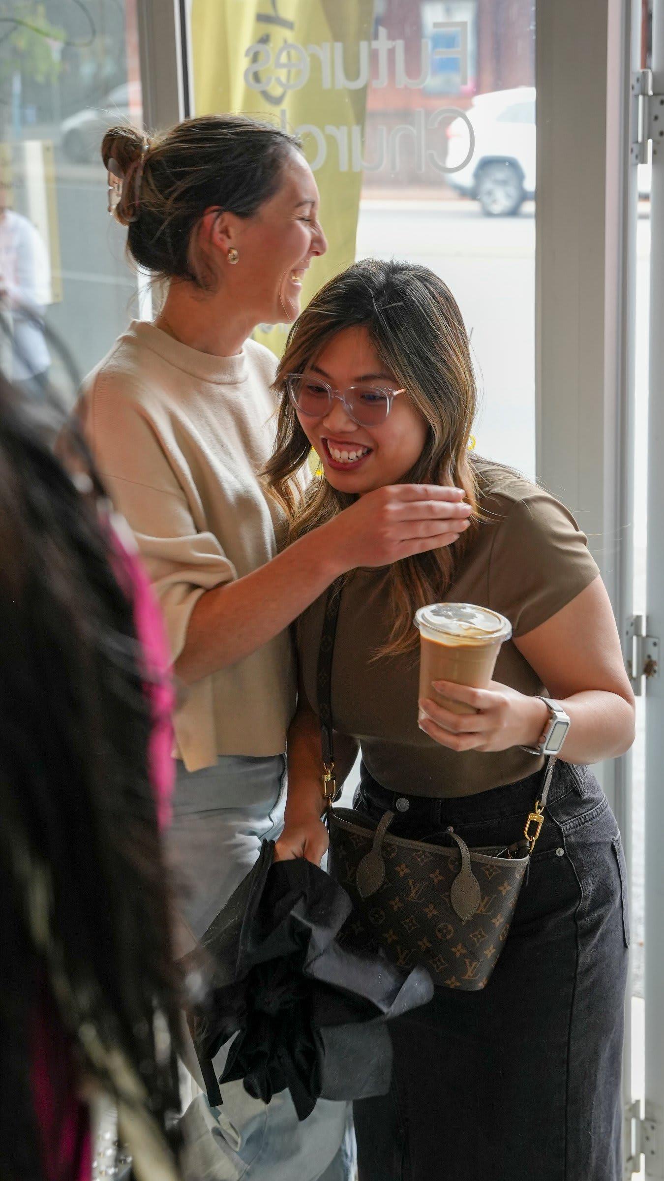 Two friends doubled over laughing with an iced coffee at the church's front door