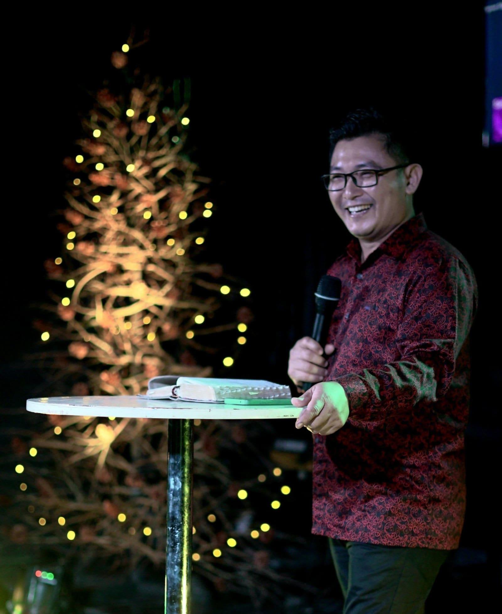 The pastor mid-laugh at the podium, Christmas lights glowing behind