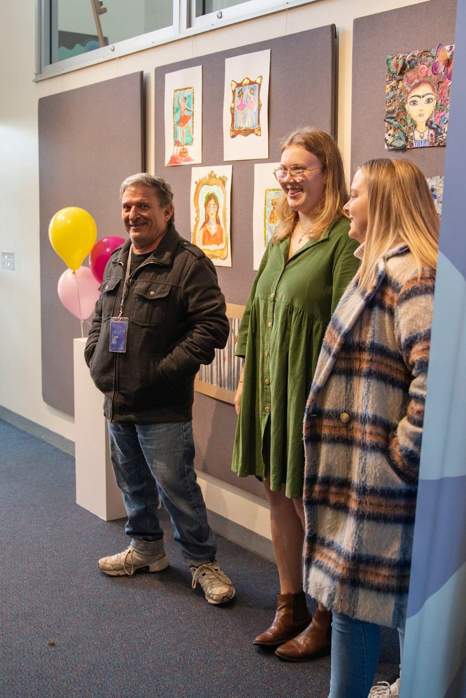 Three people smiling at the kids' art exhibition wall with balloons